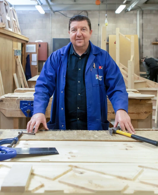 A joinery lecturer in a workshop, standing at a workbench surrounded by woodworking tools and timber, looking confidently towards the camera. A joinery lecturer in a workshop, standing at a workbench surrounded by woodworking tools and timber, looking confidently towards the camera.
